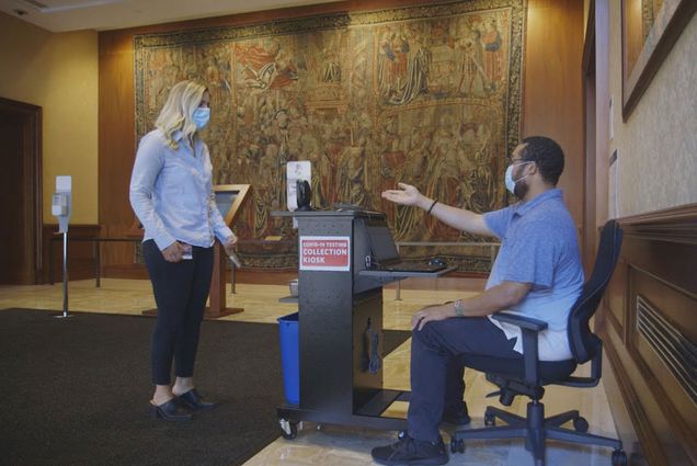 A male staff member at a COVID-19 Test Collection Kiosk prompts a woman to leave her testing swab vial in a tray on the kiosk table.