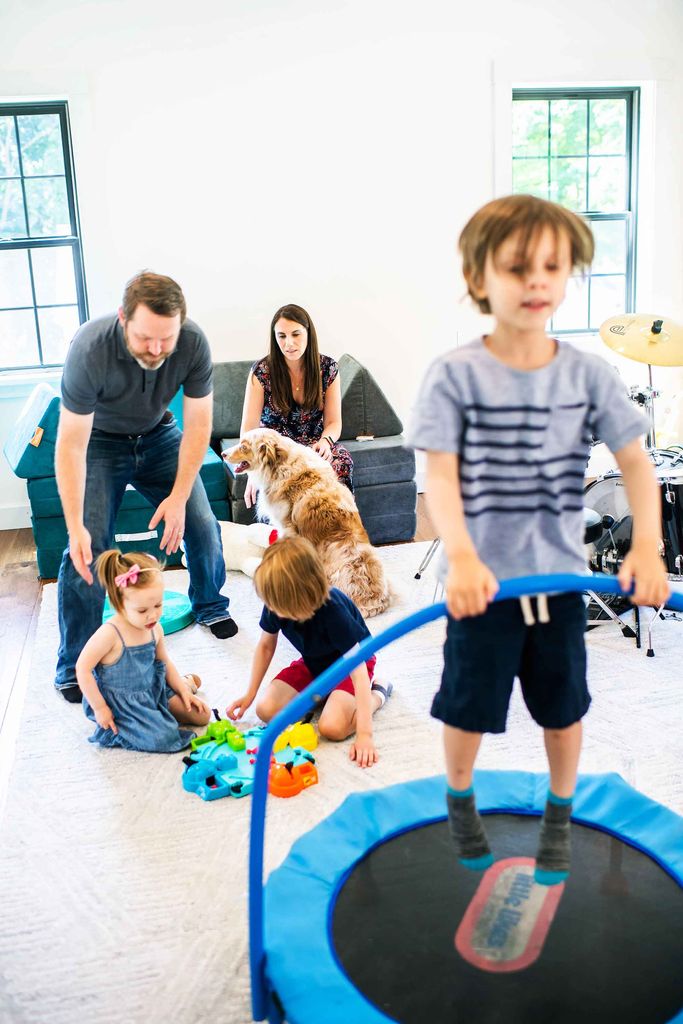 A young boy jumps on a mini trampoline while his little brother and sister play behind him as parents look on.