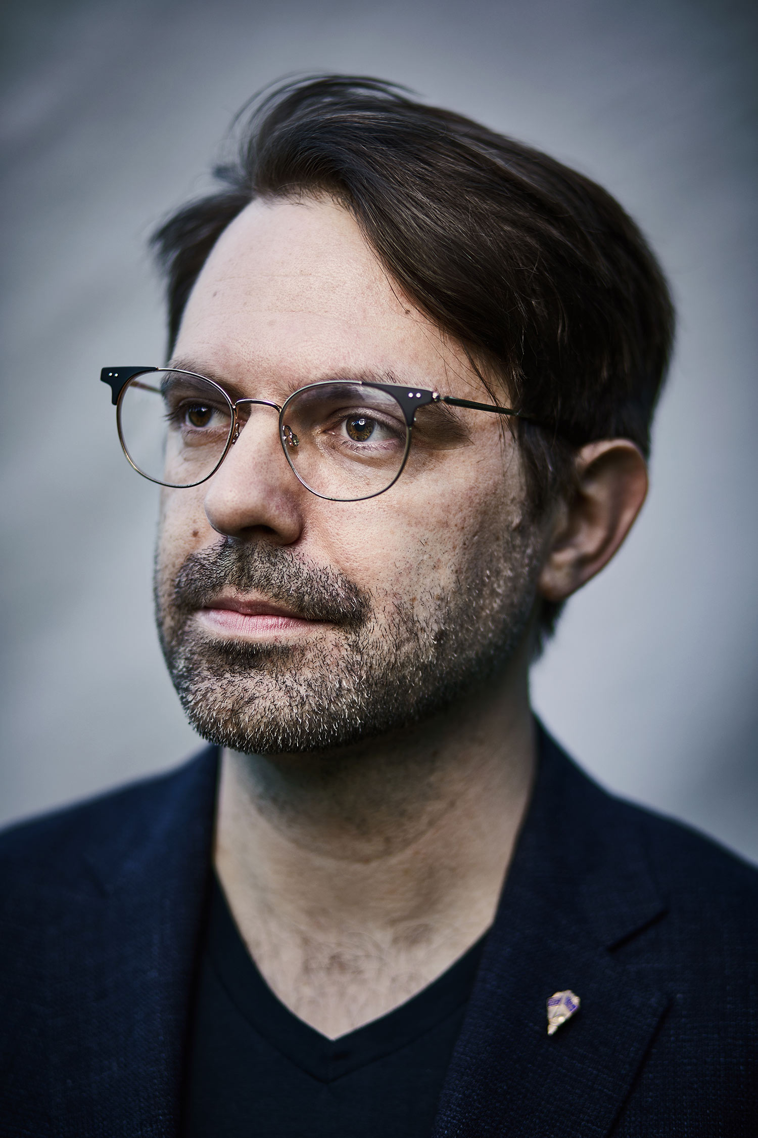 Headshot of Aaron Rasmussen, wearing glasses and a black t shirt and looking off into the middle distance on a gray background.