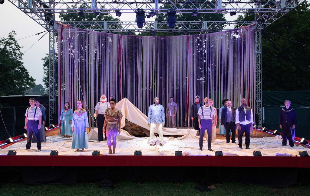 The full cast of Commonwealth Shakespeare Company’s production of The Tempest, standing on stage with their arms at their side. A backdrop made of ropes that hang is seen behind them and the stage is covered with sand.
