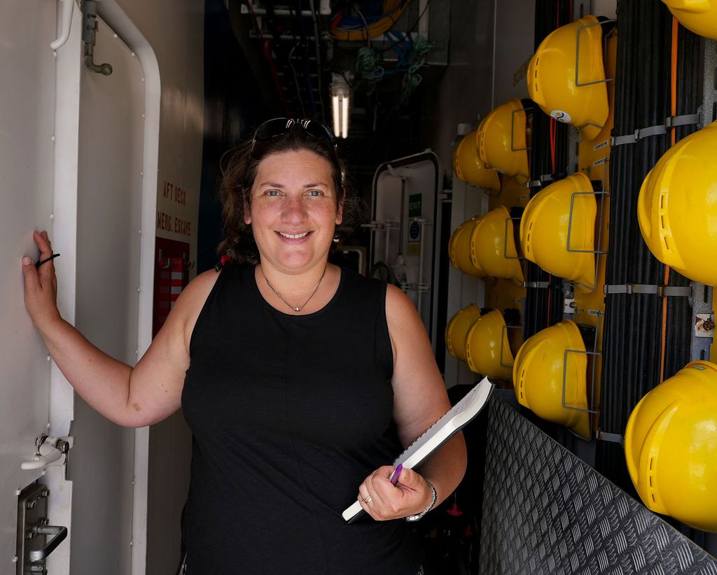 Photo of Rotjan, smiling, with her hand on the white wall of what looks like a ship hallway; yellow hard hats hang to her left and she holds a notepad.