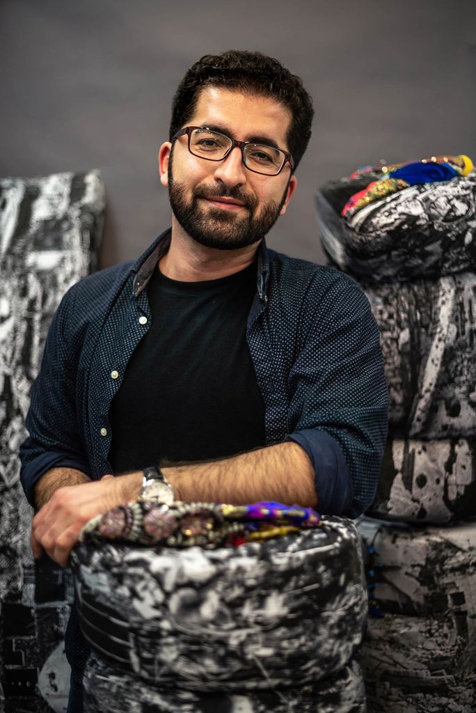 Portrait of Kamal Ahmad in a dark blue button-down smiling with his hand on what looks like a piece of his art.