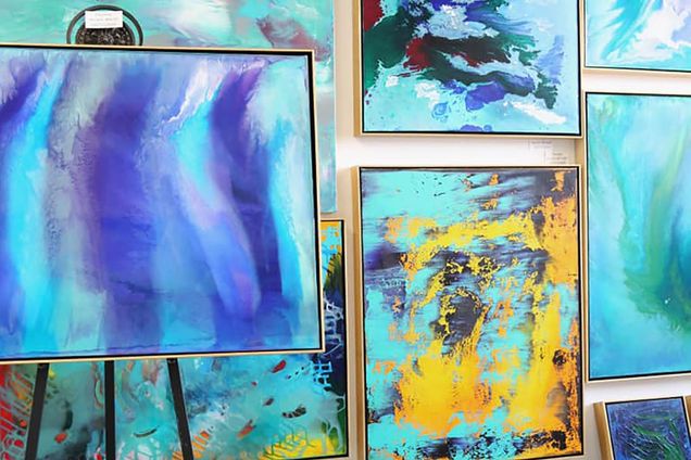 Photo of a wall of canvases painted in organic strokes of blue, teal, and gold, with an easel in the front left.