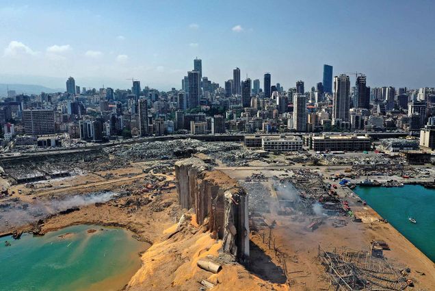 An aerial photo of the massive damage done to Beirut port’s grain silos (center) and the area around it on August 5, 2020, one day after an explosion. The yellow, orangey grain spills out of three large silos, and water pools on either side. In the background, the high-rise buildings of Beirut are seen as well as a few clouds in the sky.