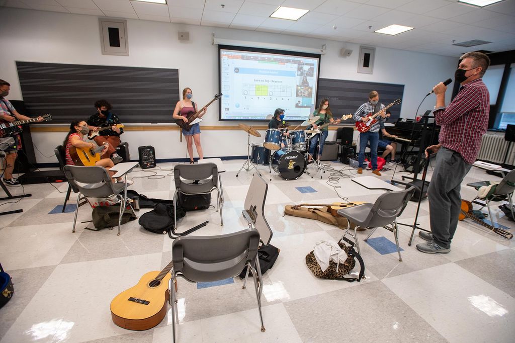 Photo of the classroom in which the rock class happens. Desks with the tabletop part folded away are arranged in a circle and some students at the front of the close hold guitars, bass guitars, and play drums. On a screen behind them, the chords to "Love on Top" by Beyonce are seen.