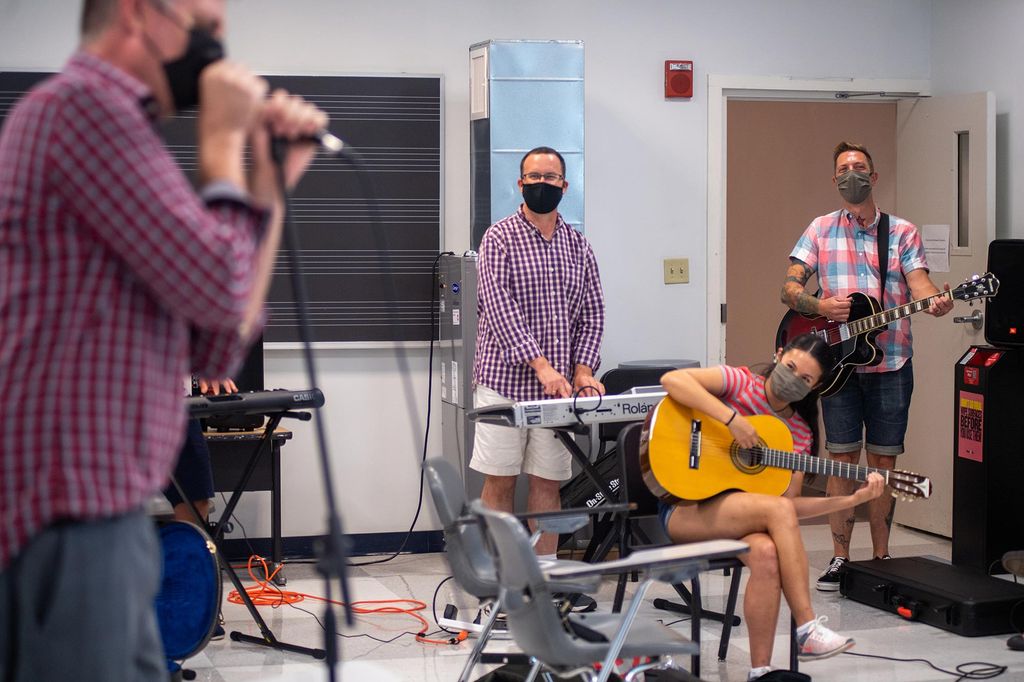 Photo of students, who appear to be smiling through their face masks, stand at a keyboard, and hold guitars and look towards Powell who has a mic in his hand and is seen blurred in the right foreground.