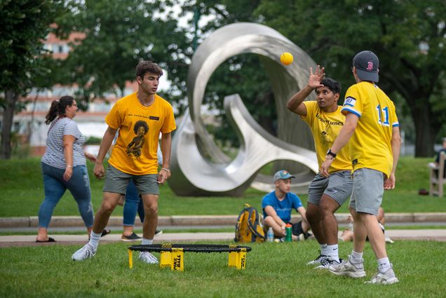 Photo of Jake Dietiker (Questrom’25), from left, Victor Verma (CAS’25), and Jeremy Gay (COM’25), playing spike ball on BU Beach August 30. They all wear bright yellow shirts and Verma reaches up to spike the small yellow ball as the others look on, Behind them, students sit in the grass or walk by.