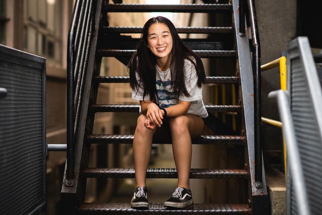 Photo of Angela Tsai with a white t-shirt with fish on it and black Vans sitting on a black staircase that may be a fire escape. She smiles wide and has long, dark hair.