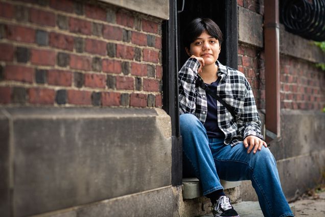 Photo of Ash Vasquez (CFA’25), sitting on a step between two brick walls. His hand is on his face, and he wears a plaid shirt and jeans.