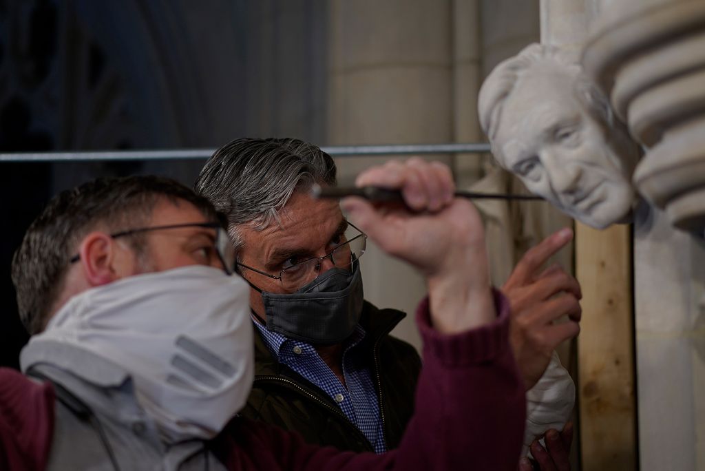 Photo of Artist Chas Fagan (right) works with stone carver Sean Callahan on his sculpture of Wiesel. Callahan holds some sort of pick and works on the bust. Fagan points towards the head. Both men wear glasses and face masks.