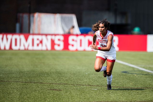 Photo of Geovanna Pereira (Sargent’23), in her white BU uniform with the number 30 on it, running across a turf field. Behind her, a blurred sign at the edge of the field reads “Women’s Soccer”