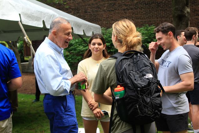 Students (from left to right) Sarah Herpst, Olivia Voelkel, and Matthew Agri speak with Colonel (Retired) T. L. Dilliplane, Ph. D. during the ROTC barbecue. Dilliplane wears a light blue button down and bright blue pants, and the student in the front wears a black backpack. A tent is seen in the background.