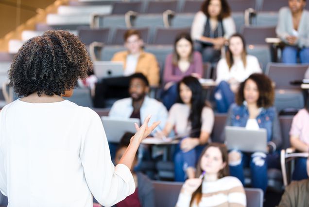 Photo of a youngish, Black woman, with curly almost shoulder length hair, wearing a white shirt, teaching at the front of a lecture hall. She is seen from behind.