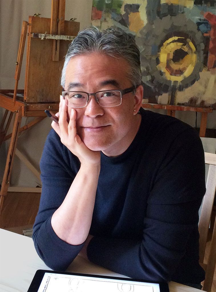 a photo of artist and author Aaron Caycedo-Kimura as he looks at camera, sitting with chin in hand, in his studio. A few of his art pieces are on display behind him.
