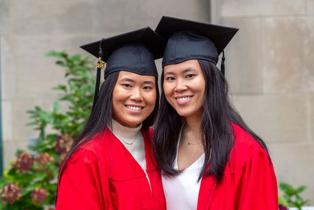 Photo of Grace Golder, (CAS’20), left, and Claire Golder (CAS’20) before commencement on October 3. The sisters smile, and wear their black caps and red gowns, with white blouses underneath. The both have longer-than shoulder length dark hair.