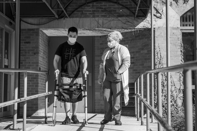 Black and white photo of Aracelis Cruz, PCA, accompanies Ethan Wang, at left, to the car after an occupational therapy session at Spaulding Cambridge Sept 29. Wang uses a walker. Both wear face masks as they exit the brick building by ramp.