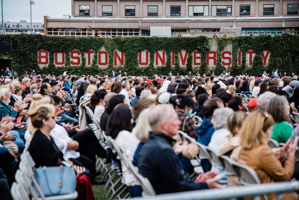 A view over the heads of a crowd of specators at BU's 147th Commencement ceremony. In the background is the Boston University sign on the wall surrounding Nickerson Field.