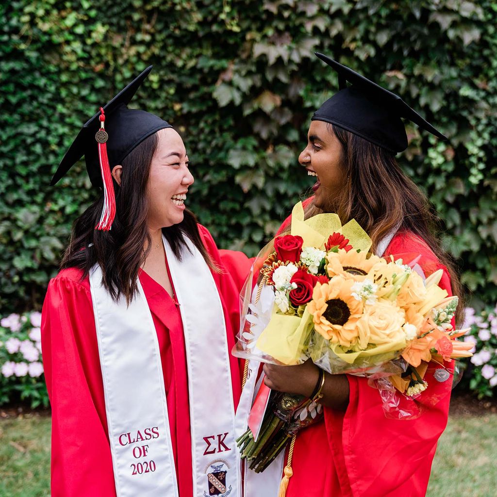 Two BU Class of 2020 graduates laugh following Commencement. One is carrying a bouquet of flowers.