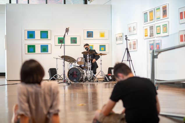 photo of Gareth Dylan Smith, Assistant Professor of Music, Music Education, playing the drums in the Stone Gallery on October 15. On the white walls behind him are colorful squares that are apart of Steve Locke’s exhibition in the gallery. Two students sit on the floor in the foreground has he plays. Gareth wears a suit and black face mask.