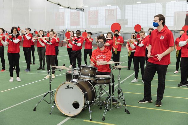 photo of members of BU's pep band, dressed in red, standing behind drummer Gabe Klavans and percussionist Thomas Lawton and playing. Most of the brass section has red mutes on their instruments.