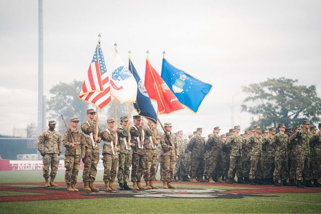 Photo of Pass-in-Review on Nickerson Field. From left to right, Commander Cadet Nathan Tadigiri, Rifleman- Cadet Kenneth Ziniti (Army), National Colors- Cadet Salem Adda-Berkane, Army Colors- Cadet Joseph Carey, Navy Colors- MIDN Sasha Wong Marine Corps, Colors-MIDN Ian Benitez-Rio Air Force Colors, Cadet Jacob Bresnahan Rifleman, Cadet Ju Young Kang (Army). They each wear camouflage uniforms and hold flags and rifles. Behind them, a larger group of ROTC members are seen.
