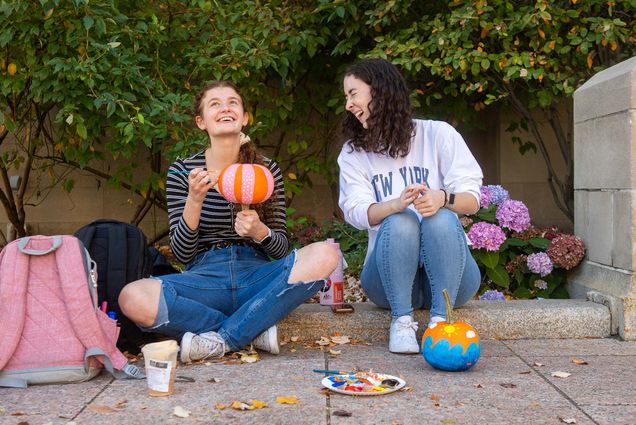 Photo of Emma Neary (Wheelock’23), left, smiling, as Grace Sullivan (COM’23), laughs while painting pumpkins in Marsh Plaza October 20. Neary wears jeans with holes at the knees and holds a pumpkin upside down as she paints pink stripes with white dots. Sullivan wears jeans and a white new york sweatshirt and sits on the curb in front of her pumpkin which is painted with blue waves.