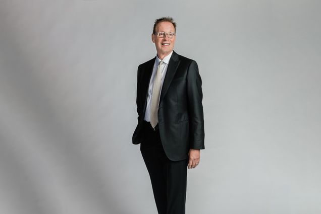 a studio photo of Boston University's Board of Trustees member Richard C. Shipley. He looks to the distance wearing a black suit in front of a grey background.