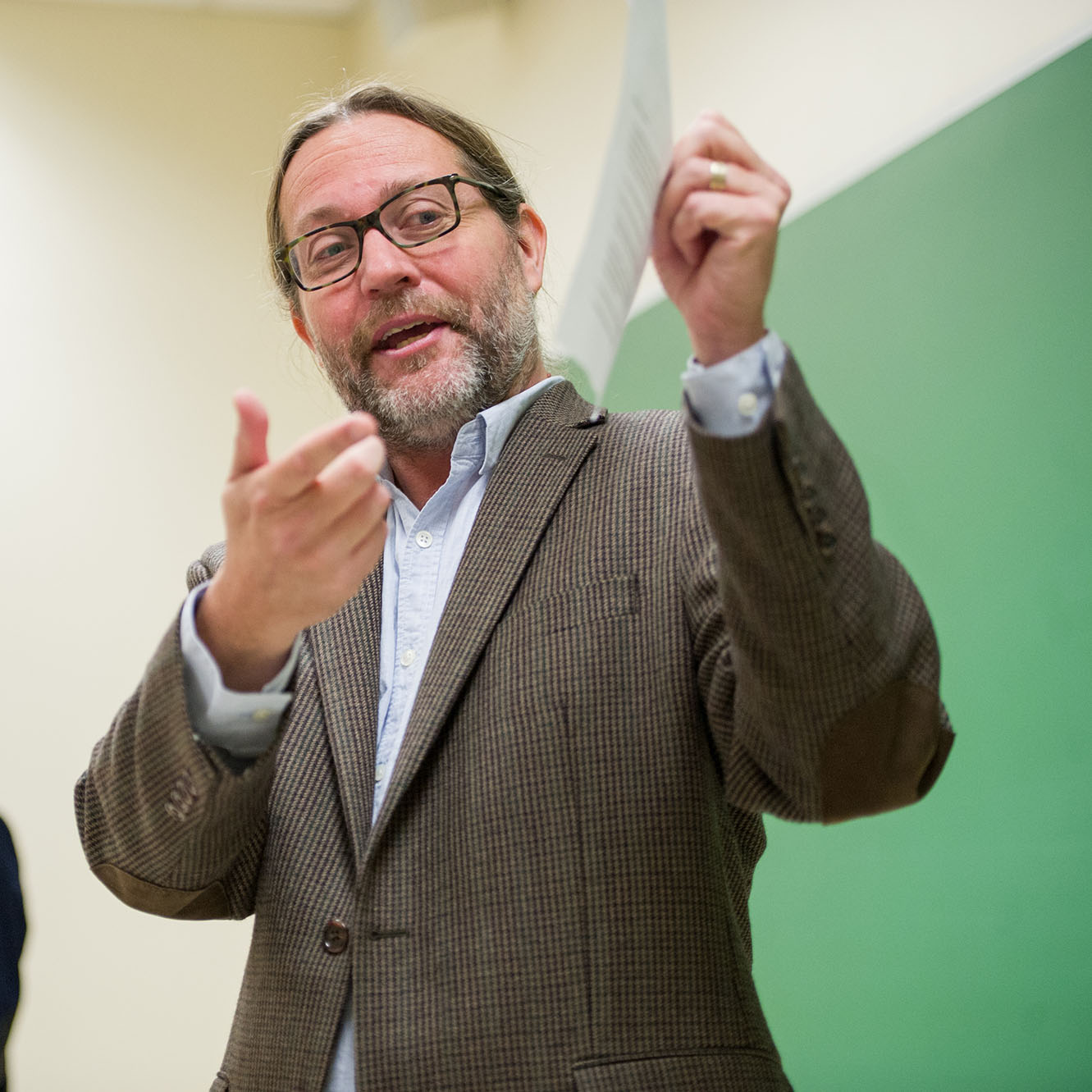 a candid photo of Kevin Barents, a CAS senior lecturer, teaching a course on Bob Dylan "Music and Words". He wears a brown blazer and gestures towards a piece of paper he's holding up.