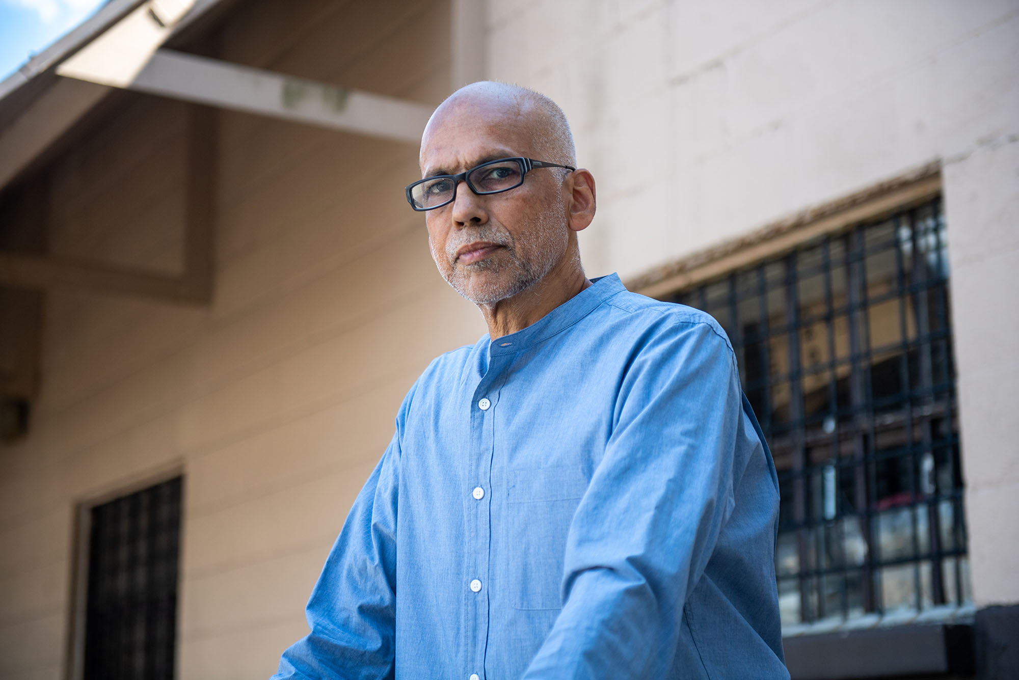 photo of André de Quadros, choir director and teacher of music in the BU prison education program, looking at the camera with a stoic expression. He wears black, rectangular glasses and a light blue collared shirt.