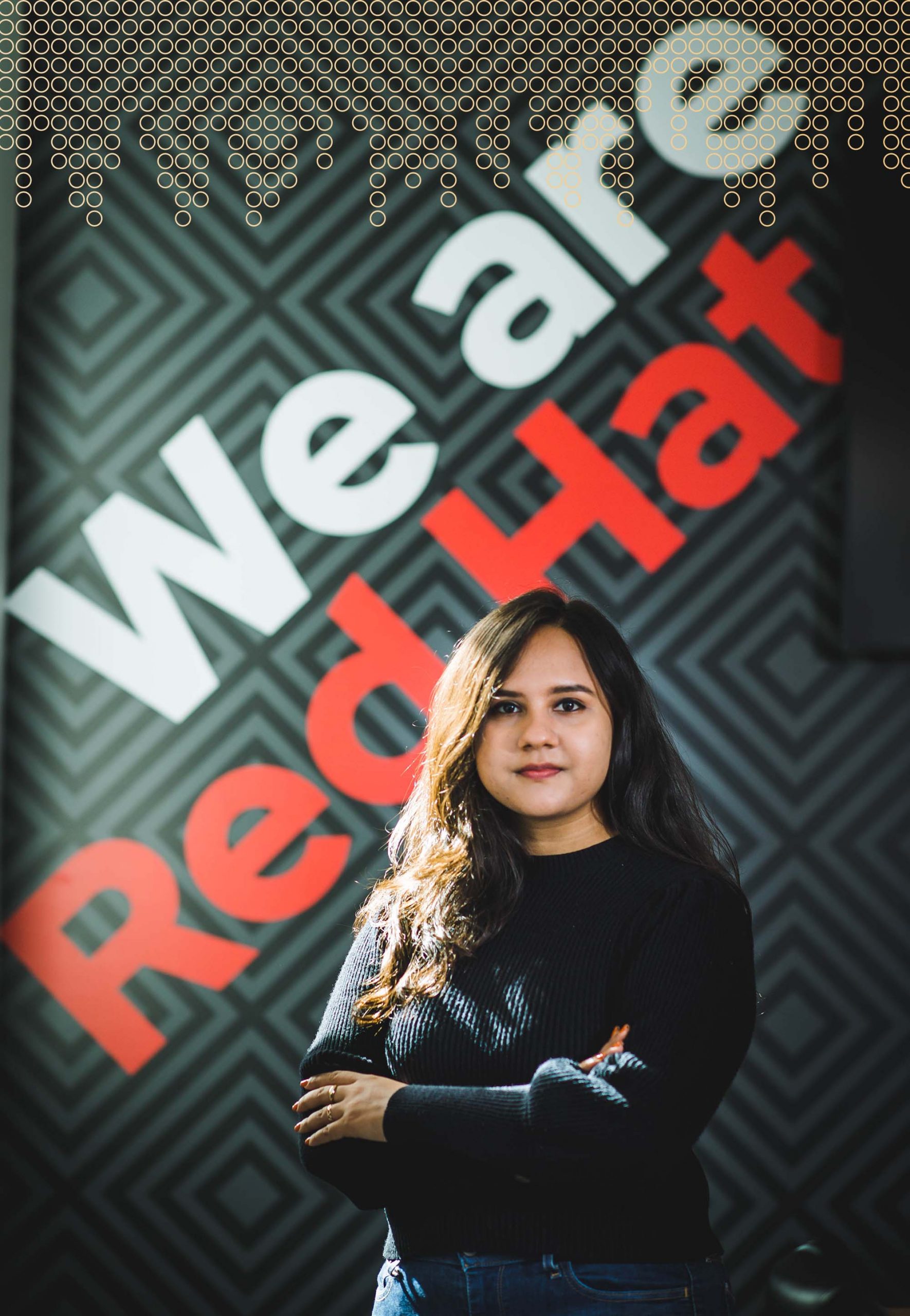 photo of Oindrilla Chatterjee. Chatterjee wears a black sweater with arms crossed and calm facial expression. The background features a dark wall witht he words "We are Red Hat" in black and white.