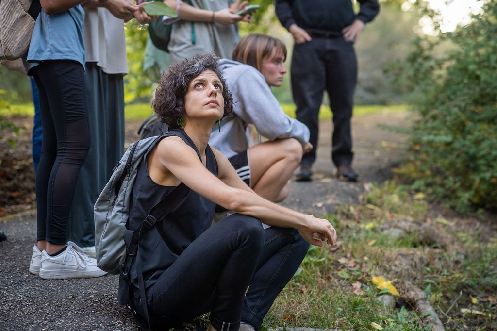 photo of Anthropologist Caterina Scaramelli, taking in the Arboretum’s mid-fall glory with her students. She wears all black and squats low as she gazes up around her as various students stand and squat beside her.