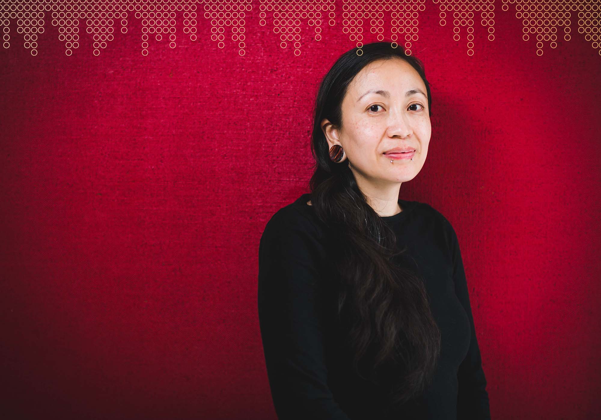 photo of Dawn Graham. They stand in front of a dark red background facing the right with a soft smile on their face. They wears snake bite piercings and a black shirt.