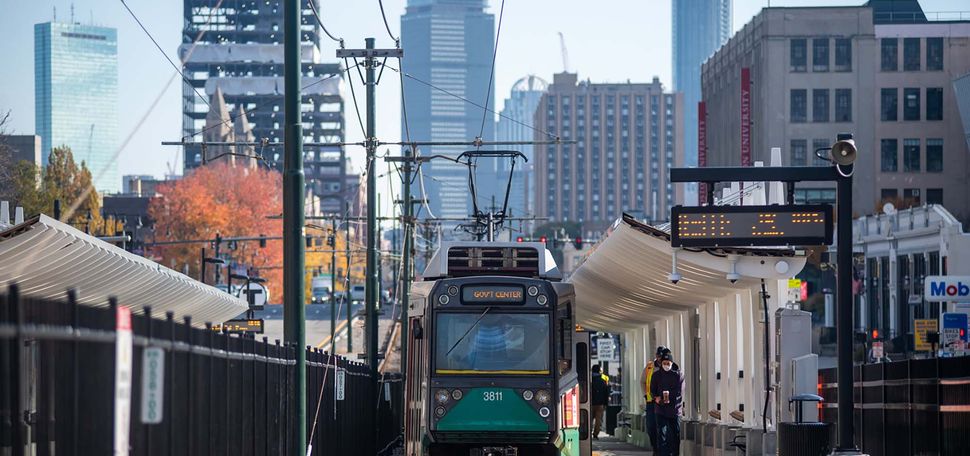 photo of an eastbound Green Line trolley pulling into the MBTA’s new Amory Street station. Two people wait under the new overhang of the station as the green trolley pulls up on the tracks.