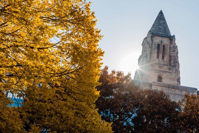 Photo of the top of an old church building. It is surrounded by Red and Yellow fall leaves as a sun glare peeks from around the top.