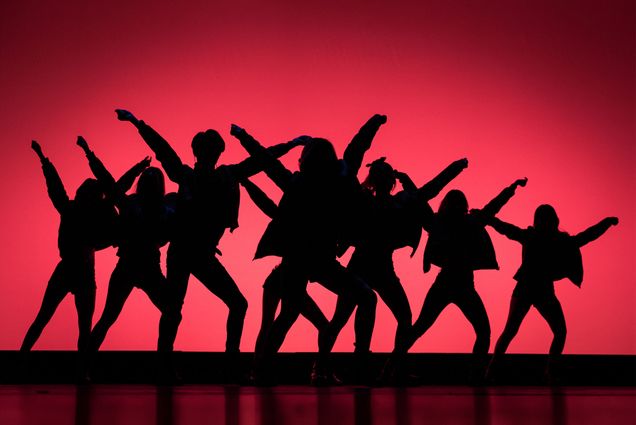 multiple kpop dancers pose with their arms above their heads in front of a red background on stage