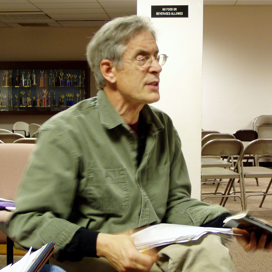 Candid photo of award-winning poet Christopher Bursk teaching a class. He wears an olive-green shirt and holds a book in hand as he speaks to his class
