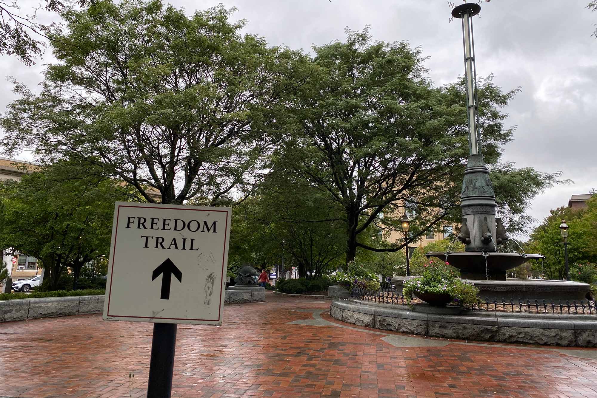 A white sign reads "Freedom Trail" with an arrow pointed at a park with green trees and red bricks