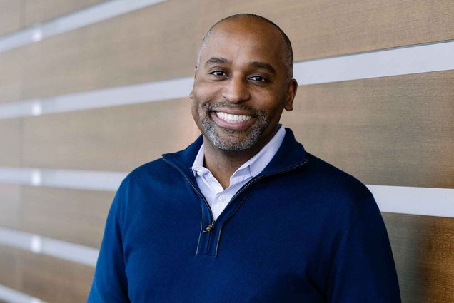 Portrait of Harvey Young, a middle-aged Black man. He wears a blue quarter zip sweater and smiles. He leans against a wall with beams running parallel to the ground.