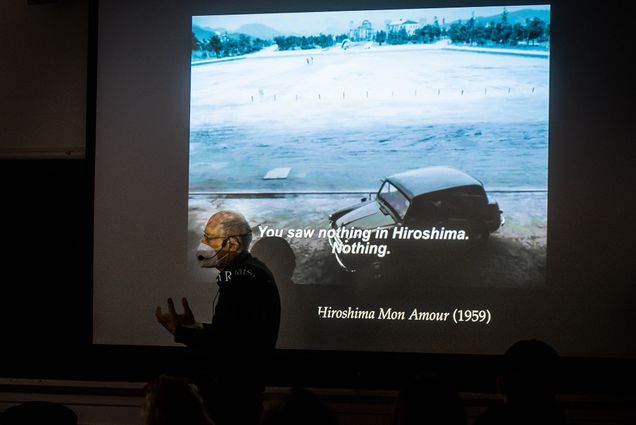 Photo of James Schmidt, wearing a white face mask, and side-lit by a projector, as he lectures, with a clip from the classic 1959 movie Hiroshima Mon Amour projected on a screen behind him.