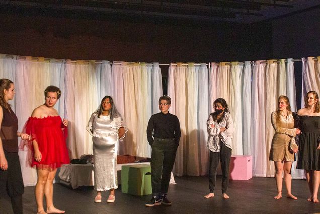 Photo of a rehearsal featuring: fEmma Permé (CAS'22) as Demetrius, Ian Aquino (COM'23) as Helena, Anoushka Thakur (CAS’23) as Hippolyta (she also plays Titania), Elizabeth Kostina (CAS'24) as Theseus, Gayane Kaligian (CAS'22) as Egeus, Carolyn (Carrie) Welter (CAS'22) as Hermia, and Ella Moran (CAS'25) as Lysander. They all wear shiny clothing, and rainbow pastel-colored curtains hang behind them.