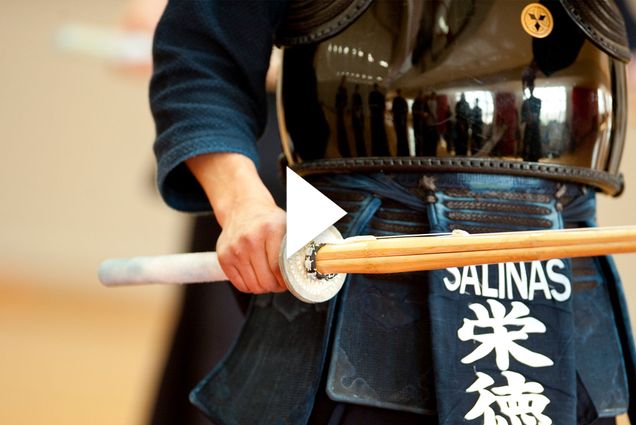 photo of a person in a kendo stance, holding a long wooden stick, and wearing a navy uniform and black reflective chest protector. White triangle play button overlays image.