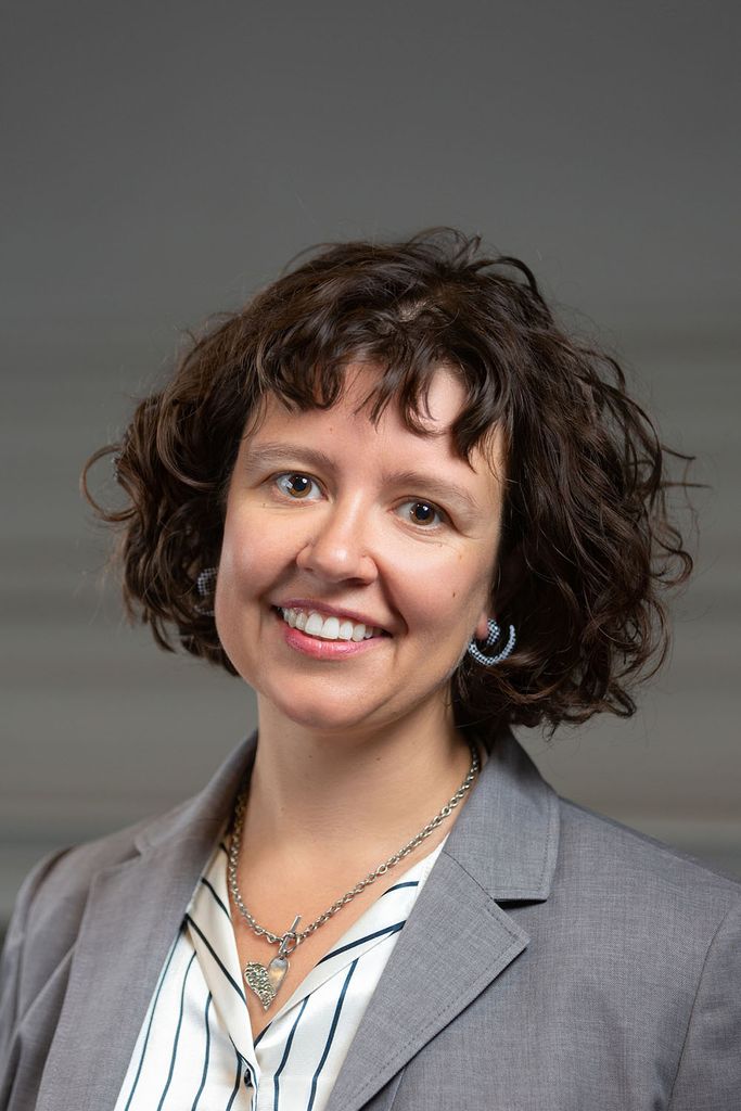 Headshot of Romy Ruukel. She has short, wavy brown hair, and smiles. She wears a gray blazer.