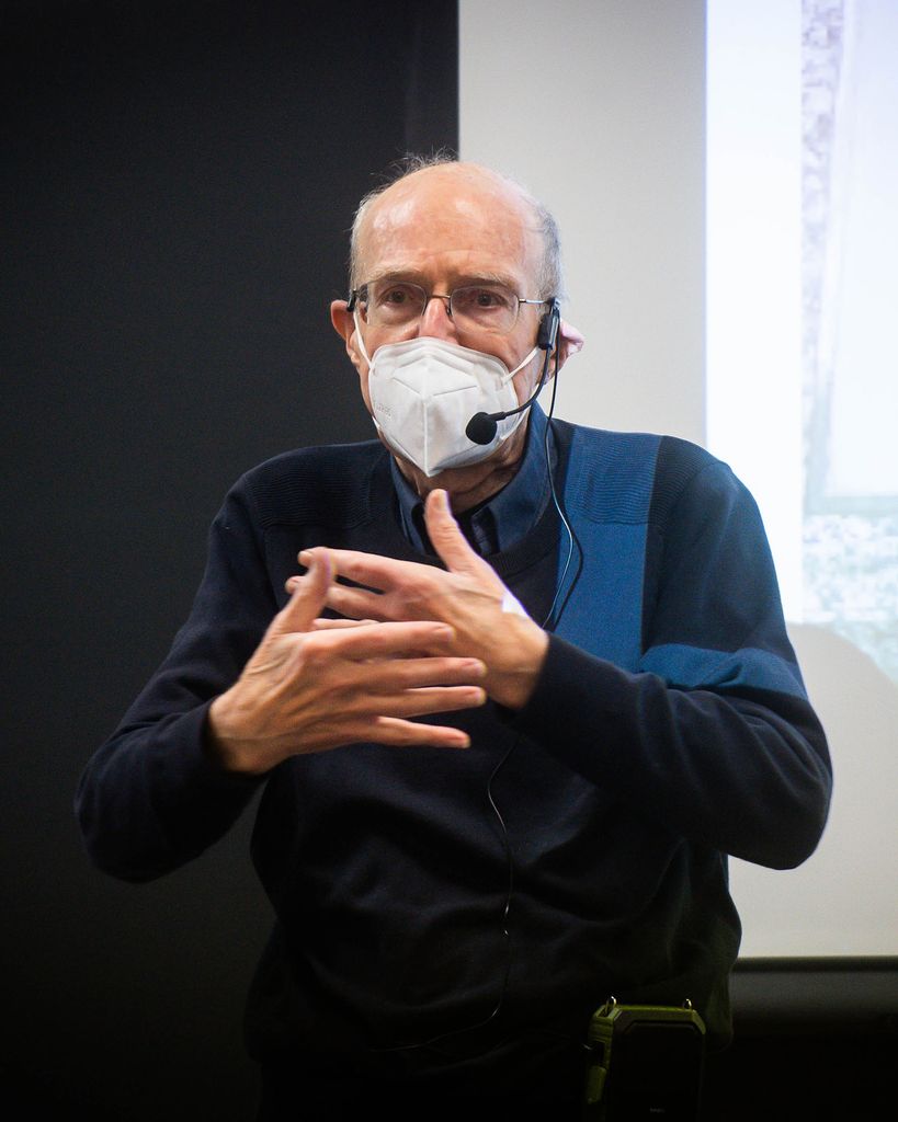 Photo of James Schmidt, who wears glasses, a white face mask, and a blue sweater, gesturing with his hands pointing towards one another, as he lecture for his class on Catastrophe and memory.