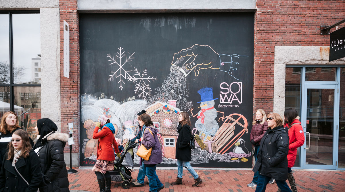 Photo of a group of people dressed in winter coats walking by a wall decorated with winter-y drawings in chalk, including a snowman, snow, and a sled.