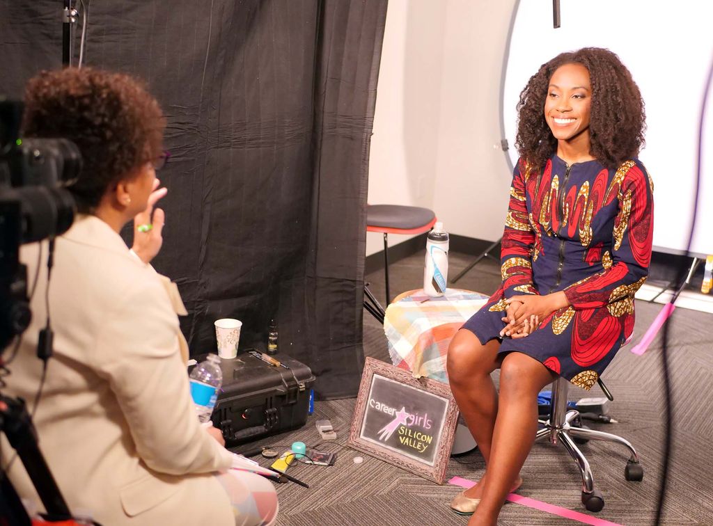 Photo of Calhoun interviewing a young Black woman, on camera, who wears a blue and red dress and sits in front of a white screen.