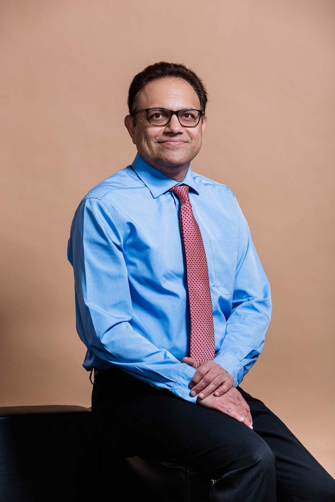 Portrait of Arun Upneja, dean of SHA, wearing a blue button down and a red tie. He smiles, wears glasses with black frames and sits with his hands folded in his lap.