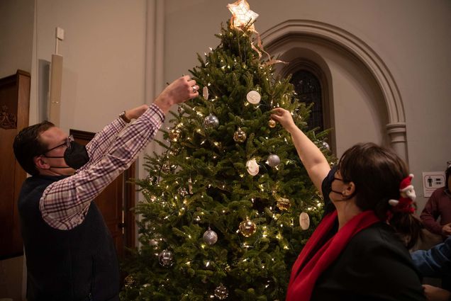 Photo of Clayton McCleskey (STH’24), left, and Sharon Solomon. (CAS’14) put the finishing touches on the Marsh Chapel Christmas tree. Both wear face masks. The tree is decorated with yellow lights and cream-colored ornaments, and has a five-pointed star on top.