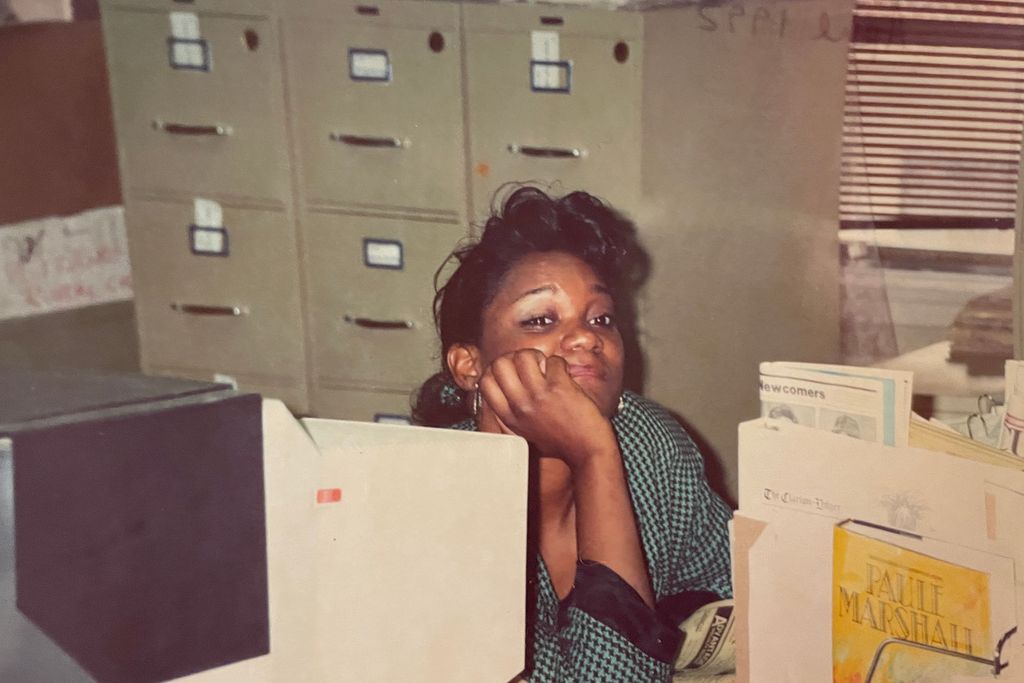 Photo of Douglas as a young journalist. She wears a teal pull over and rests her hand on her cheek. She sits behind a desk with file cabinets in the background. The photo appears to have been taken of a printed photo.