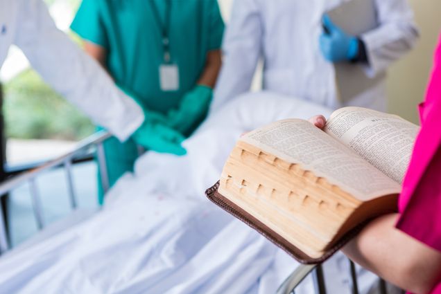 a group of doctors and nurses stand around a patient while a bible is being read. Photo by iStock/SDI Productions