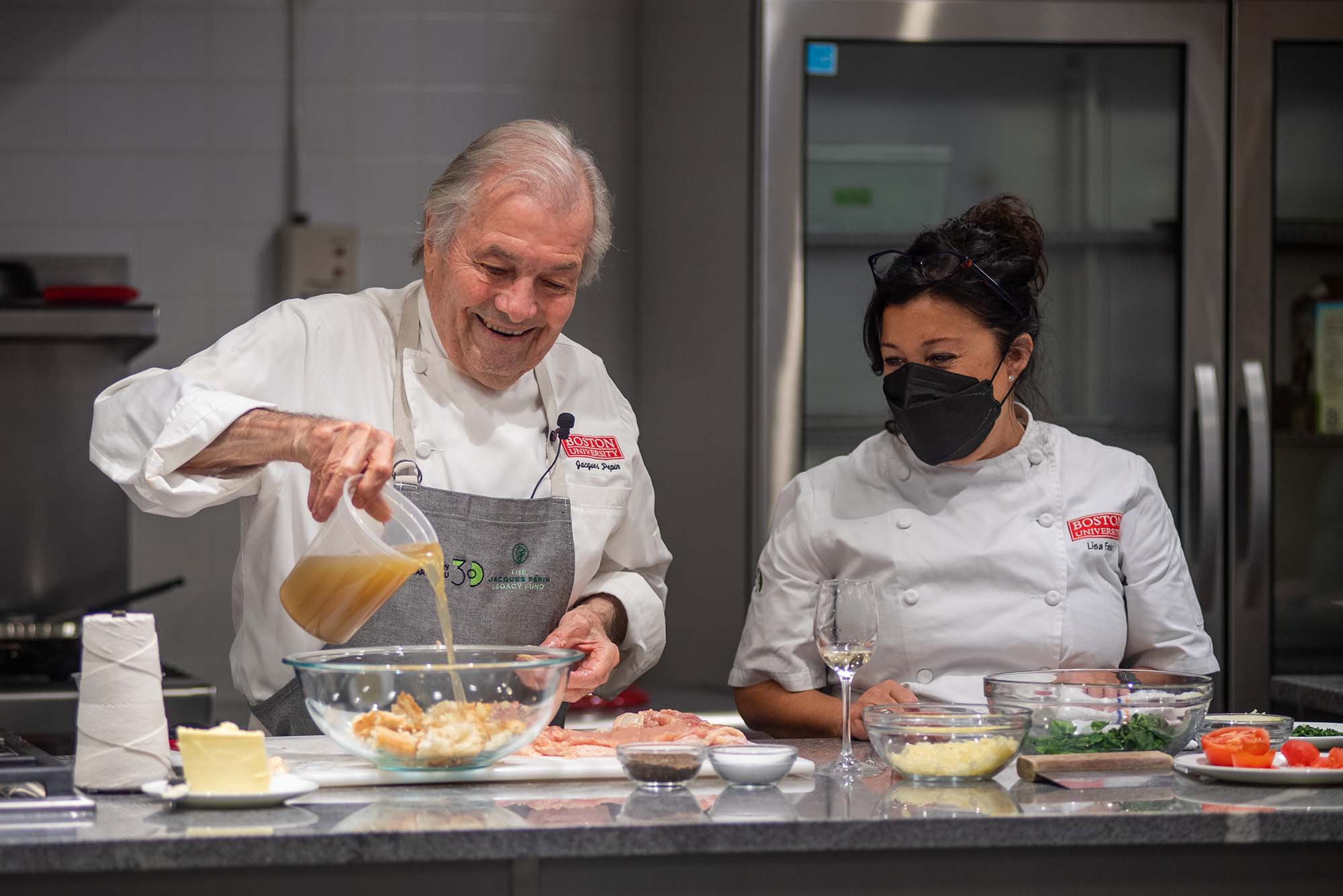 Photo of Jacques Pépin (Hon.’11), assisted by Lisa Falso-Doherty, as he pours stock into the bread and spinach stuffing for chicken ballottine in the newly renovated Groce-Pépin Culinary Innovation Lab kitchen. Pépin wears a gray Pépin foundation apron and chef jacket with BU logo, and Falso-Doherty also wears a chef jacket with the BU logo and wears a black mask. Bowls of ingredients rest on the counter before them.